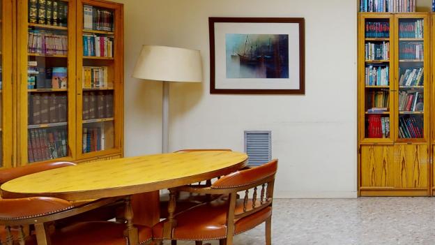 La sala cuenta con paredes de tono amarillo claro, suelo de terrazo grisáceo y mobiliario de madera que incluye varias estanterías repletas de libros, dos mesas de estudio de madera clara con sillas de cuero marrón, un cuadro en la pared y un mapa de España colgado junto a una mesa auxiliar de oficina.