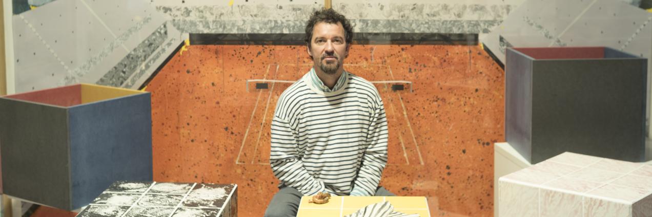Hombre sentado mirando a cámara frente a un mural donde está pintada una pista de tenis