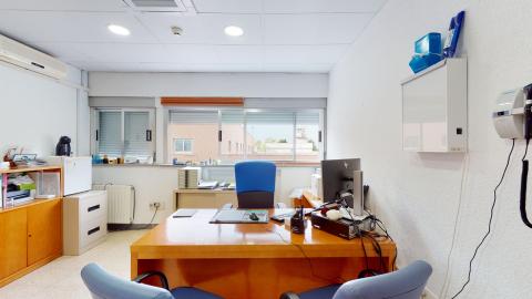 Consulta en color blanco. Al fondo hay ventanas de izquierda a derecha. Debajo de éstas, una consola. En el muro izquierdo una archivero de metal y una consola de madera. Al centro el escritorio de madera con sillas azules. En el muro derecho una lámpara para ver radiografías. 