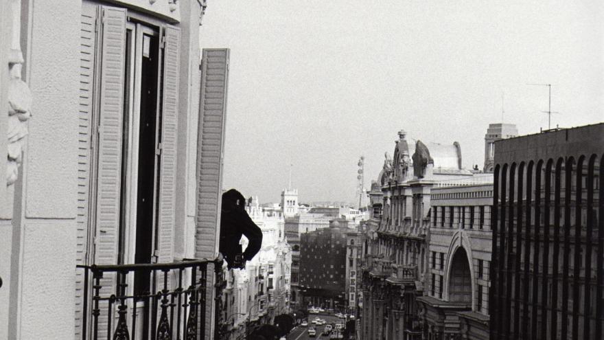 Vista urbana tomada desde lo alto de un edificio; una calle con tráfico, una persona se asoma a uno de los edificios