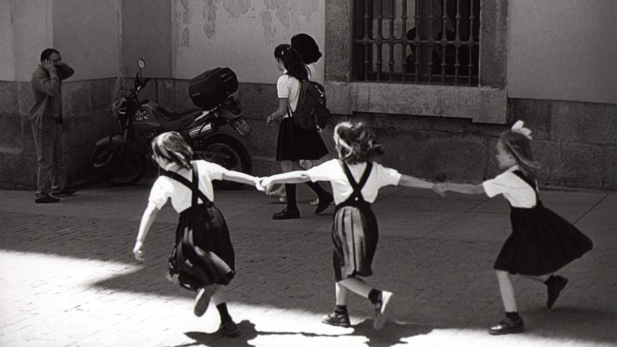 Tres niñas vestidas de uniforme, de espaldas y agarradas, juegan en la calle