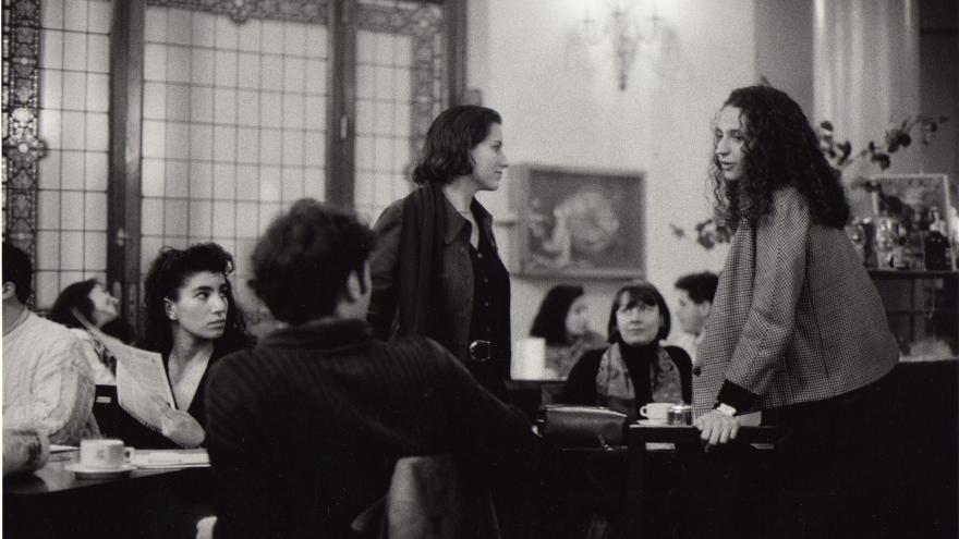 Interior de un café, varias personas sentadas en diferentes mesas, en una de las mesas dos personas están de pie y apoyadas sobre la mesa