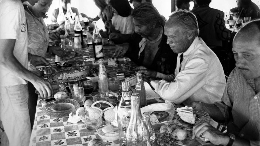 Varias personas, sentadas o de pie, comiendo alrededor de una mesa