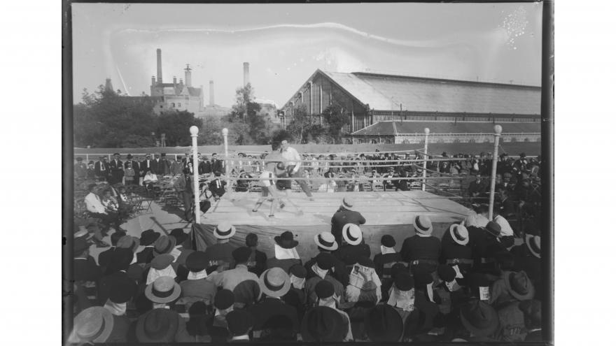 Foto en blanco y negro con un ring de boxeo y público observando el espectáculo