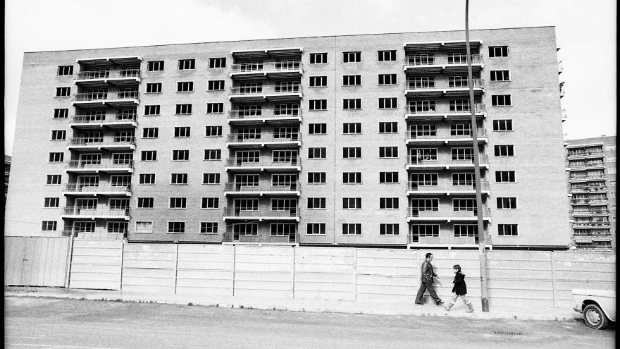 Fotografía de Javier Campano, edificio rodeado por una valla de obras, un hombre y un niño se cruzan por la acera en el barrio de Hortaleza, Madrid