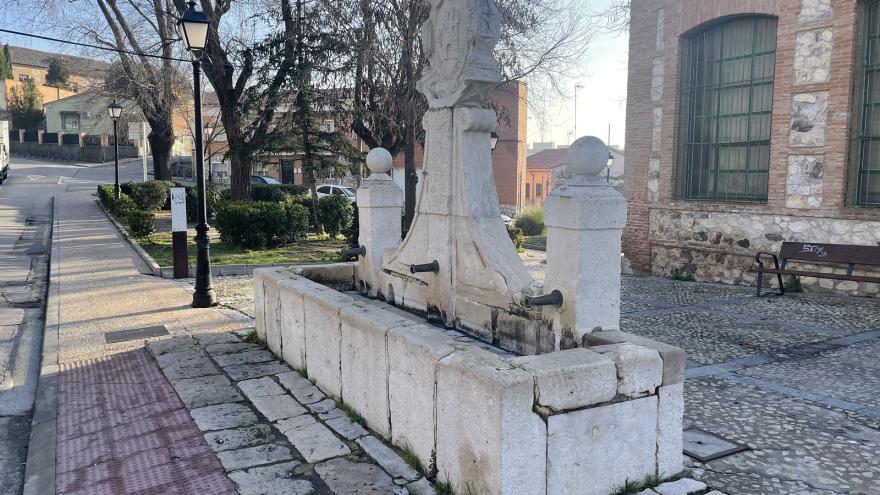 Fuente de piedra blanca con un pilón de agua, dos columnas y un escudo central con inscripciones