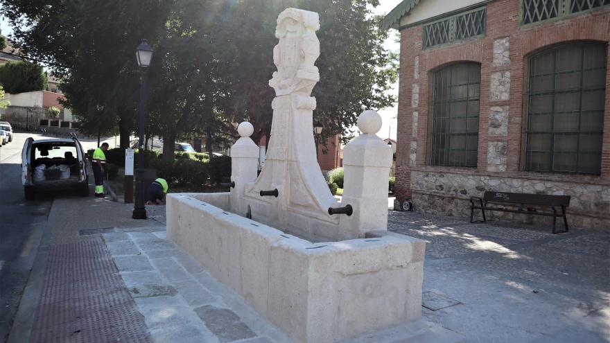 Imagen frontal de una fuente de piedra blanca con dos columnas laterales, escudo frontal con inscripciones y pilón de agua