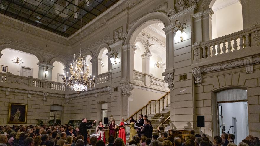 Grupo de mujeres y hombres cantando a coro en el salón de un edificio con galería y columnas