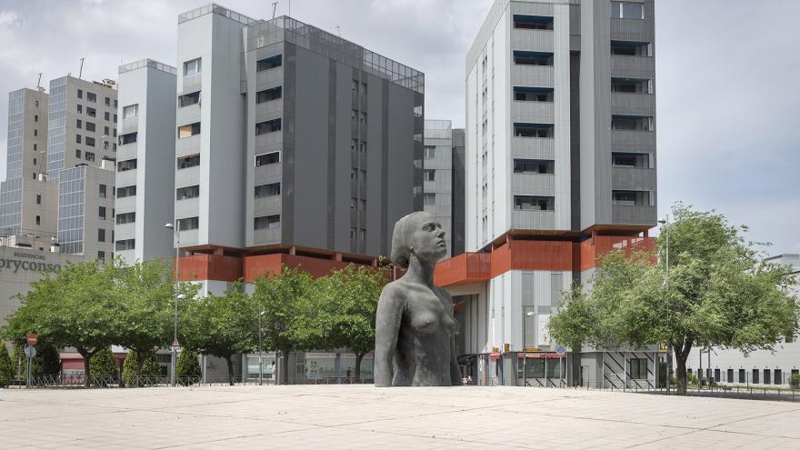 Vista urbana de Coslada, escultura de medio cuerpo de una mujer ubicada en el centro de una rotonda, al fondo varios edificios 