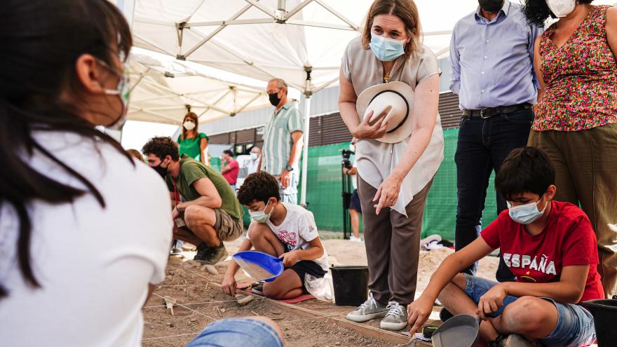 La consejera hablando con unos niños que están sentados en la arena buscando restos arqueológicos