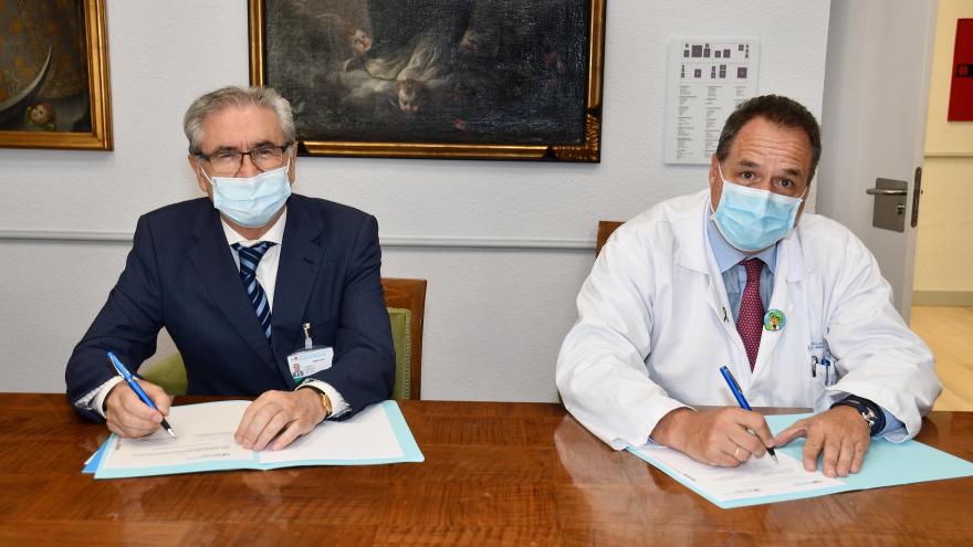 Dos hombres uno con traje y corbata y el otro con bata de médico sentados en una mesa firmando el acuerdo