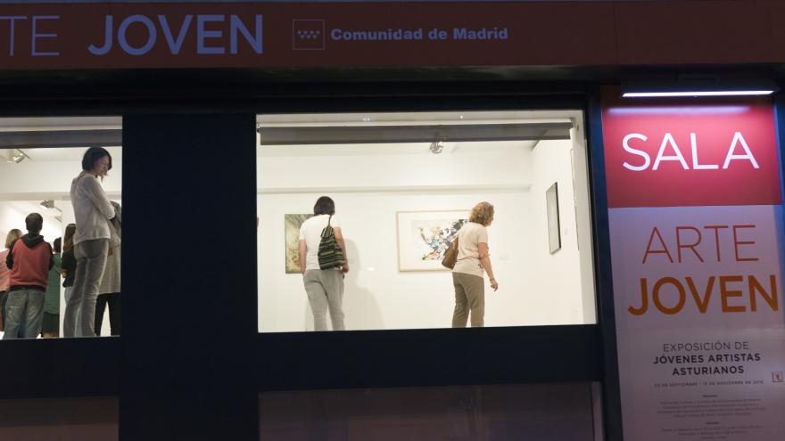 Vista desde el exterior de una estancia de la sala de arte joven iluminada, con visitantes conociendo una exposición