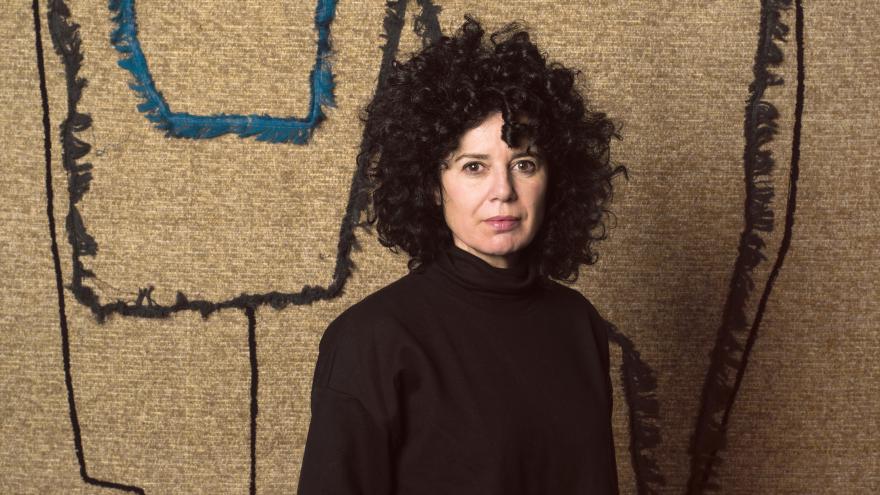 Retrato de una mujer frente a un lienzo que está compuesto por trozos de textiles, pespuntes y costuras