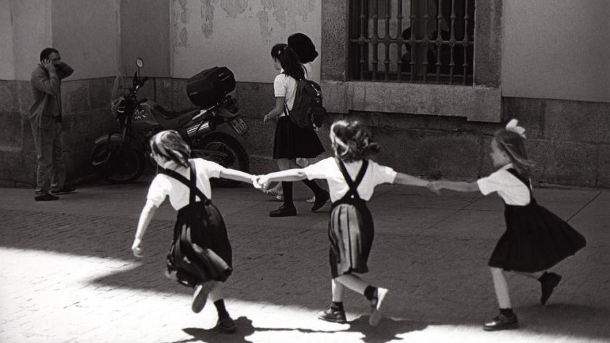 Tres niñas vestidas con uniforme, de espaldas, jugando en la calle