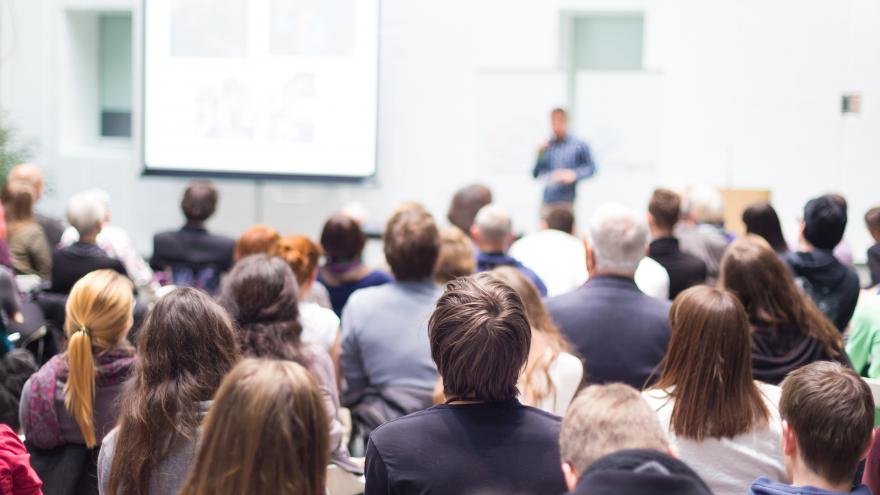 Un grupo de personas sentadas en un aula, escuchando a un conferenciante que está al fondo, delante de una pared y de una pizarra