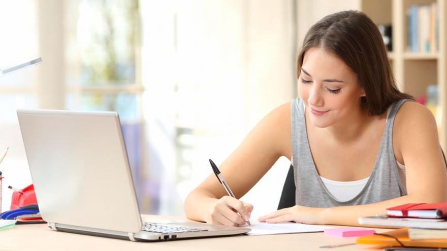 Chica escribiendo en un cuaderno con un portátil delante