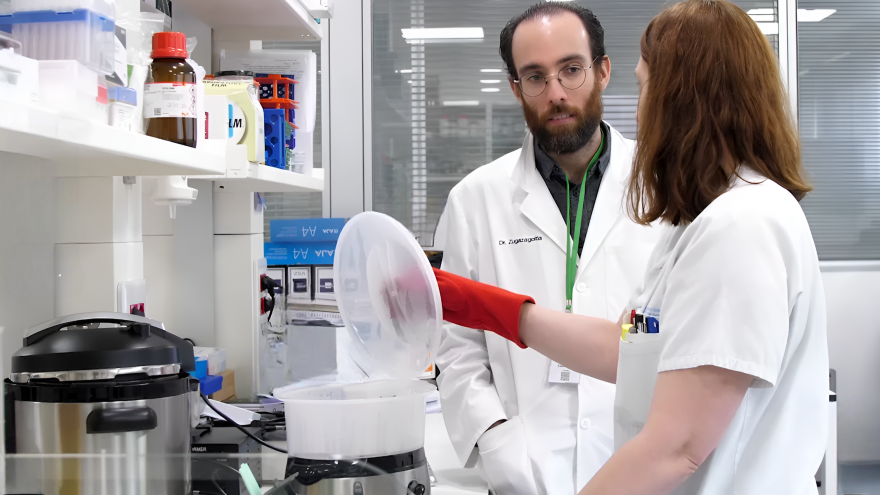 Investigador escucha a investigadora que trabaja con unos recipientes en un laboratoio