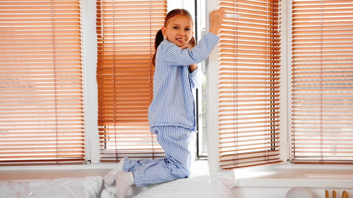 niña en pijama subida a un sofa abriendo la ventana y haciendo un gesto de silencio