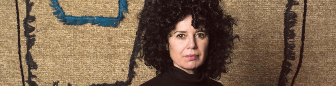 Retrato de una mujer frente a un lienzo que está compuesto por trozos de textiles, pespuntes y costuras