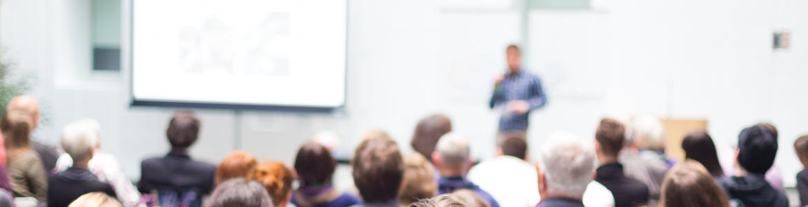 Jornadas Un grupo de personas sentadas en un aula, escuchando a un conferenciante que está al fondo, delante de una pared y de una pizarra