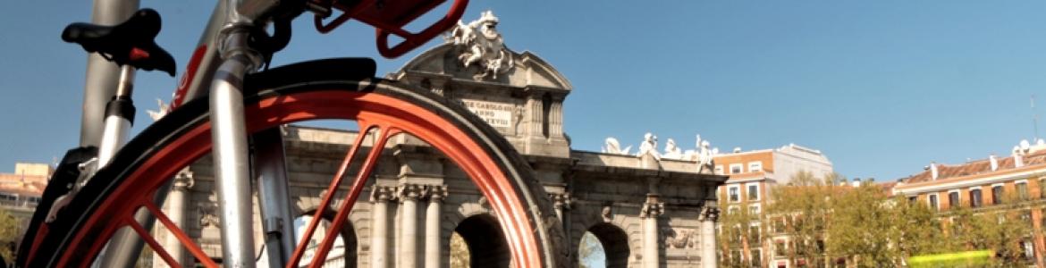 IMAGEN DE LA PUESTA DE ALCALÁ VISTA TRAS LOS RADIO DE LA RUEDA DE UNA BICICLETA ROJA