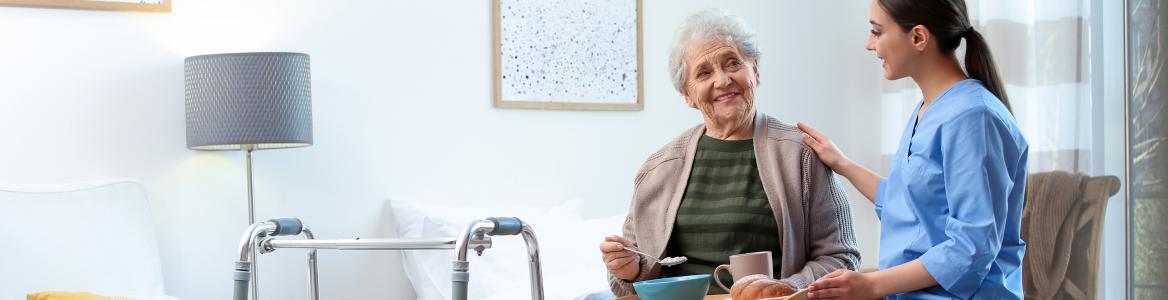 Una persona mayor en una habitación de un centro residencial comiendo acompañada de una profesional sanitaria