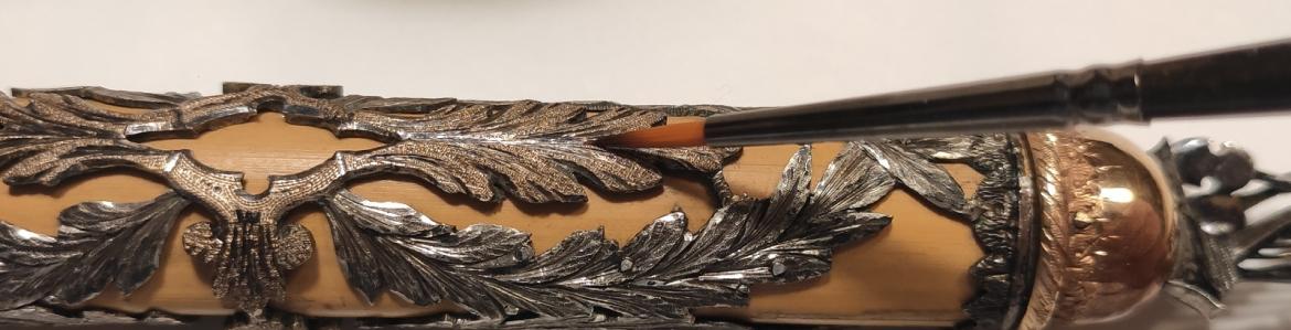 Proceso de restauración de la batuta del Real Conservatorio de Música un pincel limpia una batuta cilíndrica con decoración vegetal en plata