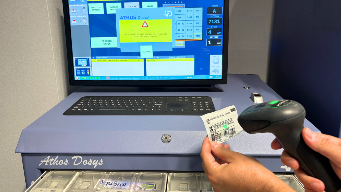 Mano con una pistola de leer códigos juntoa pantalla de ordenador con aviso de alerta y cajón debajo con medicamentos de alto riesgo