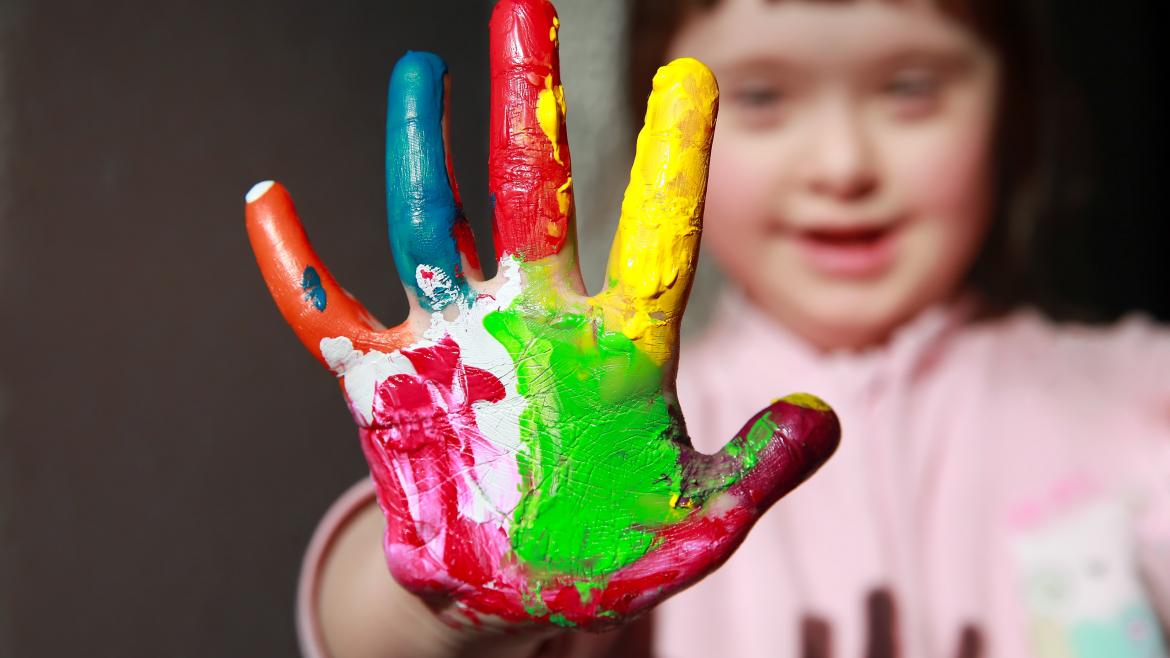 Una niña con Síndome de Down con una mano pintada de muchos colores extendida hacia la cámara