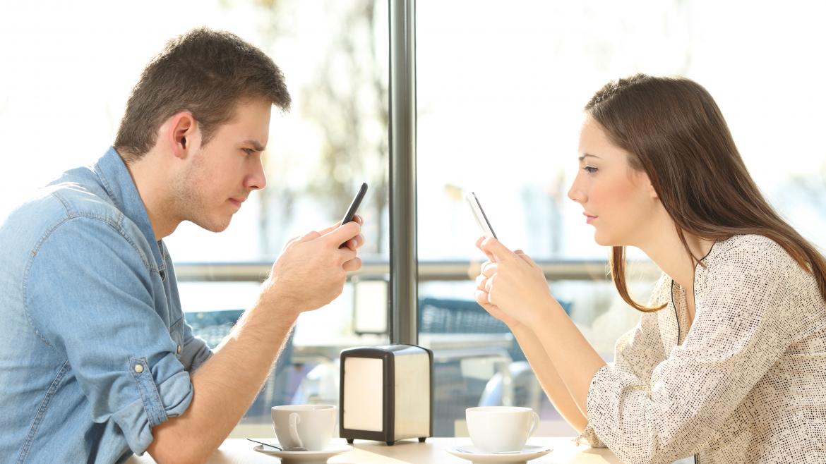 Un hombre y una mujer sentados frente a frente en una cafetería, cada uno está mirando la pantalla de su móvil