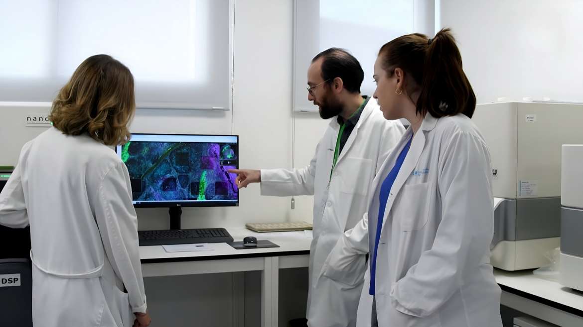 Tres personas con bata blanca, dos mujeres y un hombre trabajando en un laboratorio frente a una pantalla