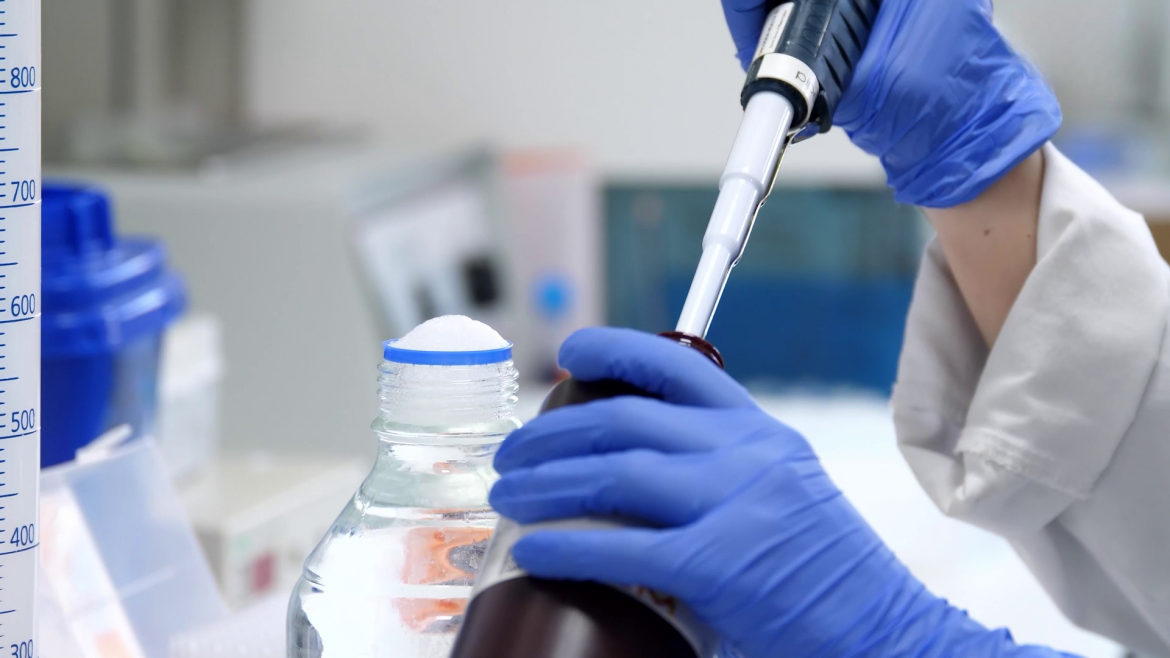 Manos de profesional con guantes azules con pipeta introduciendo líquiso en un frasco en laboratorio