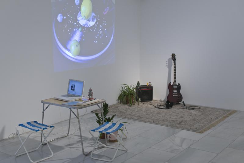 Instalación artística con piezas diversas como una mesa, un ordenador, una alfombra, una guitarra eléctrica