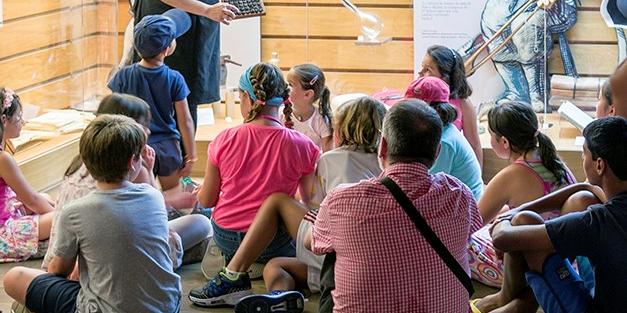 Niños y niñas en el centro de interpretación mirando a la educadora de la actividad que les explica cómo se hace el vídrio