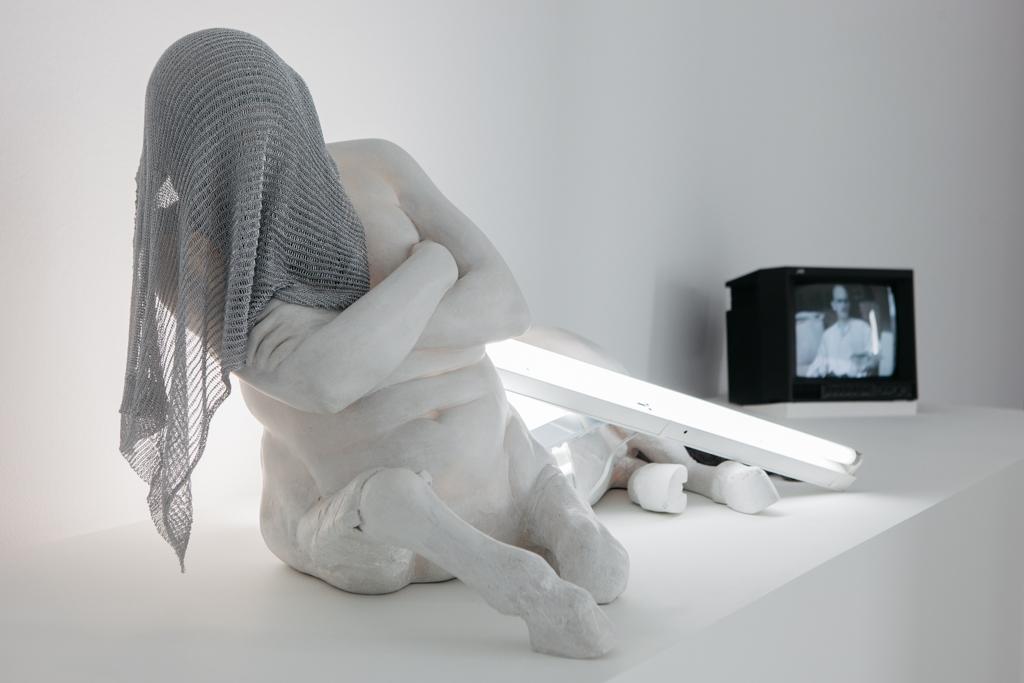 Expo Bernardí Roig. Foto Guillermo Gumiel Escultura de un fauno cubierto con una malla metálica. De fondo varios fluorescentes encendidos y amontonados y una televisión que emite un audiovisu