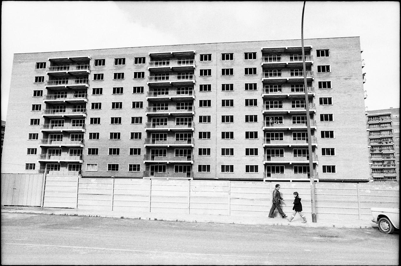 Fotografía de Javier Campano, edificio rodeado por una valla de obras, un hombre y un niño se cruzan por la acera en el barrio de Hortaleza