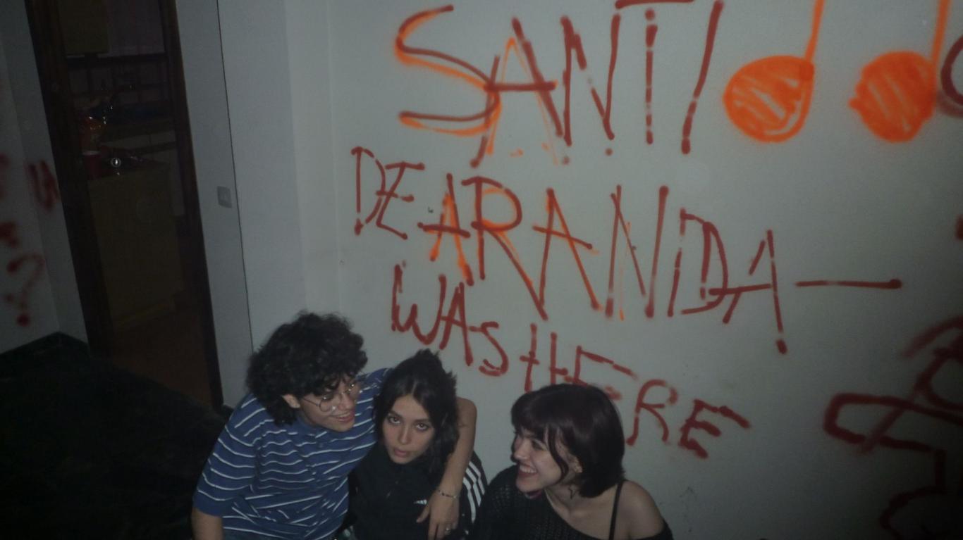 3 jóvenes sentados con el nombre de su grupo musical pintado en la pared de atras