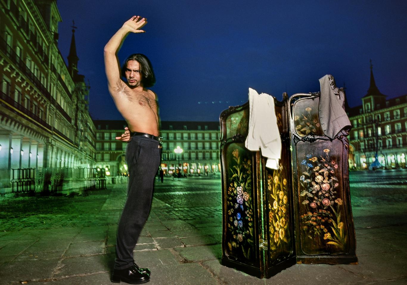 Hombre bailando flamenco en una plaza mayor; junto a él un biombo decorado con flores del que cuelgan dos prendas de vestir