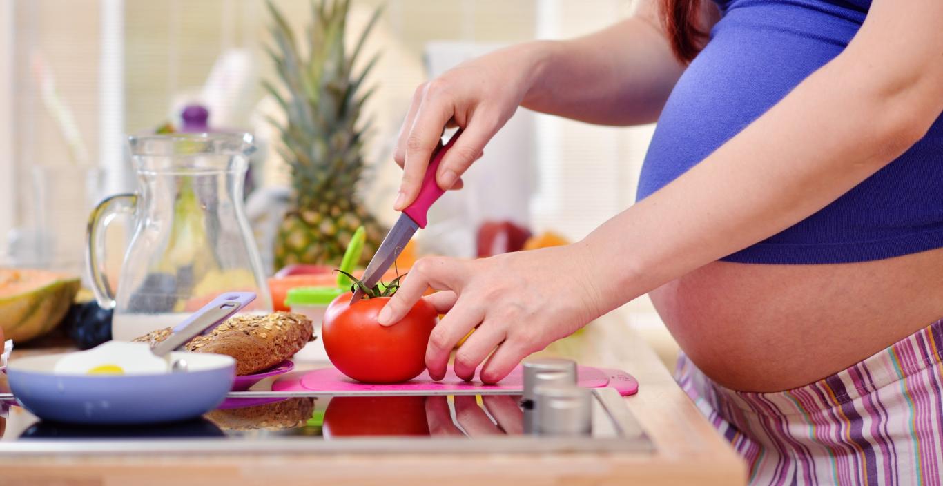 Las manos de una mujer embarazada cortan un tomate sobre la encimera de la cocina