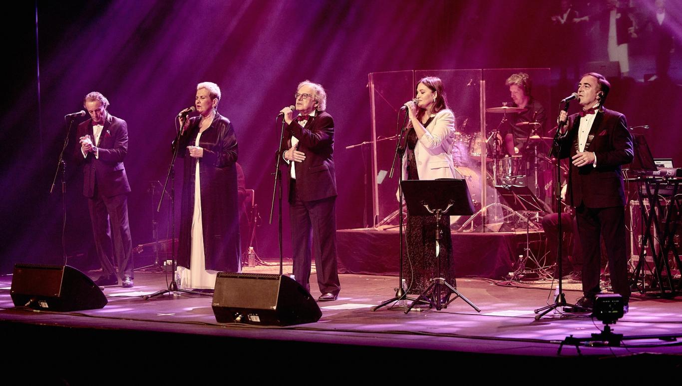 Imagen de agrupación actuando en un escenario con tres hombres y dos mujeres cantando junto a un hombre tocando la batería