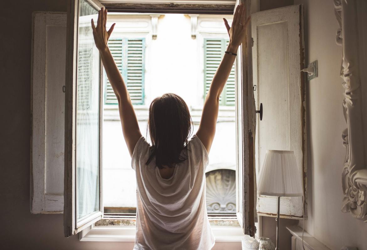 Chica de espaldas con los brazos levantados frente a una ventana de un domicilio abierta