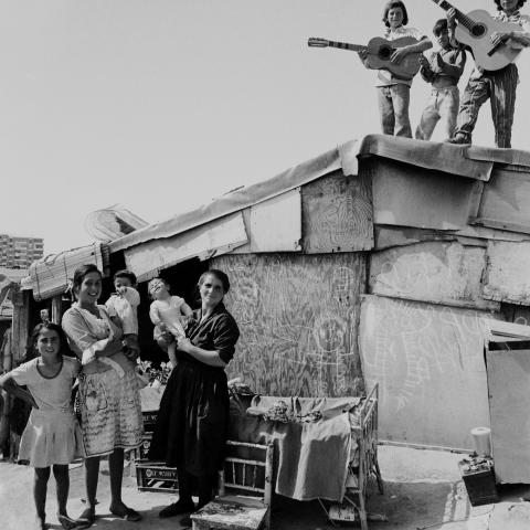 En un poblado, una niña y dos mujeres de pie con dos bebés en brazos, tres niños de pie tocando la guitarra clásica y dando palmas