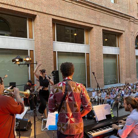 Grupo de músicos toca en un escenario con público sentado en un patio
